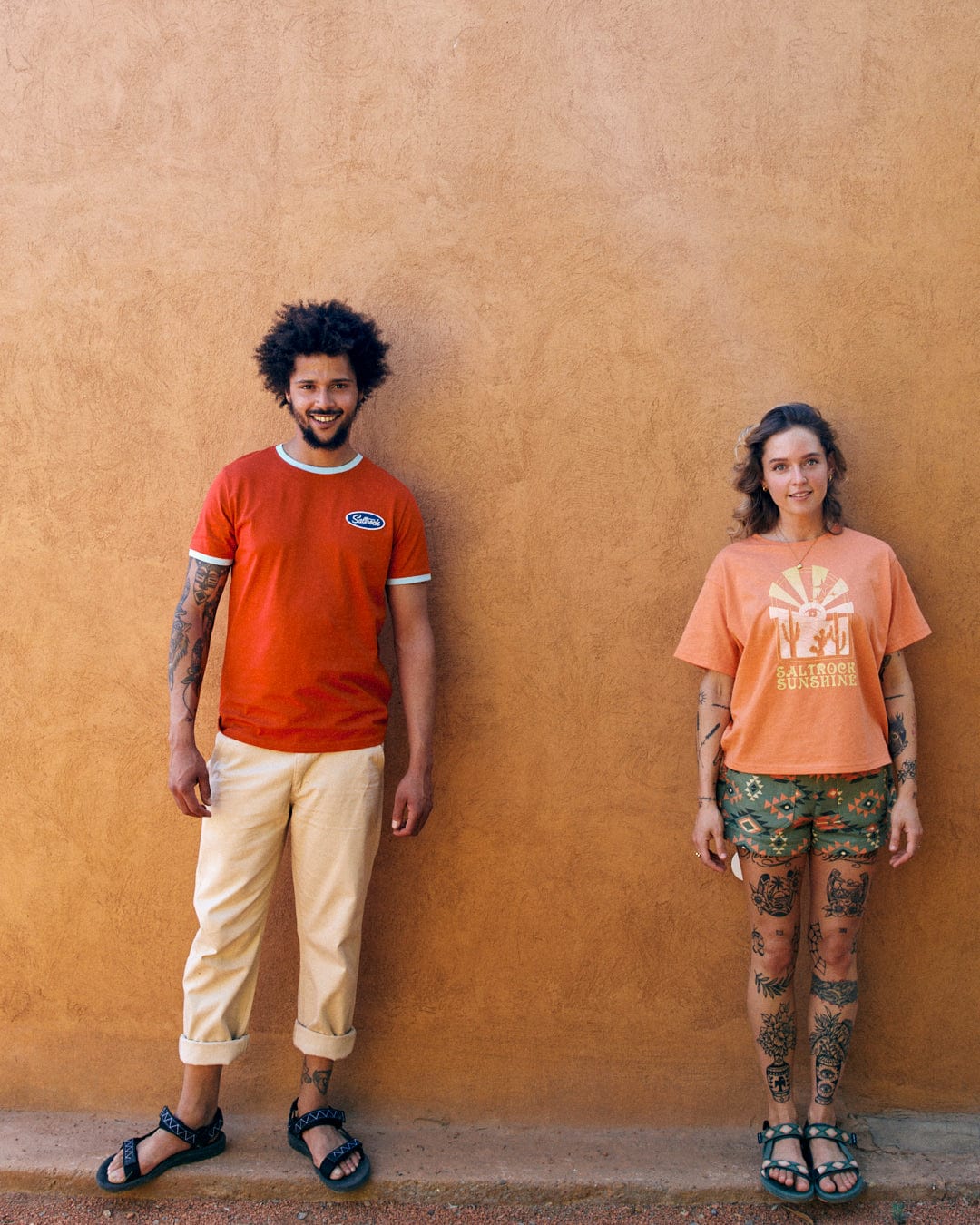 Two people stand against a beige wall. The person on the left wears a red shirt and light-colored pants, exuding Saltrock sunshine branding style. The person on the right, in a peach shirt and patterned shorts with relaxed drop shoulders, is wearing the Saltrock Sunshine - Recycled Womens Cropped T-Shirt - Orange from Saltrock, both displaying their vibrant tattoos.