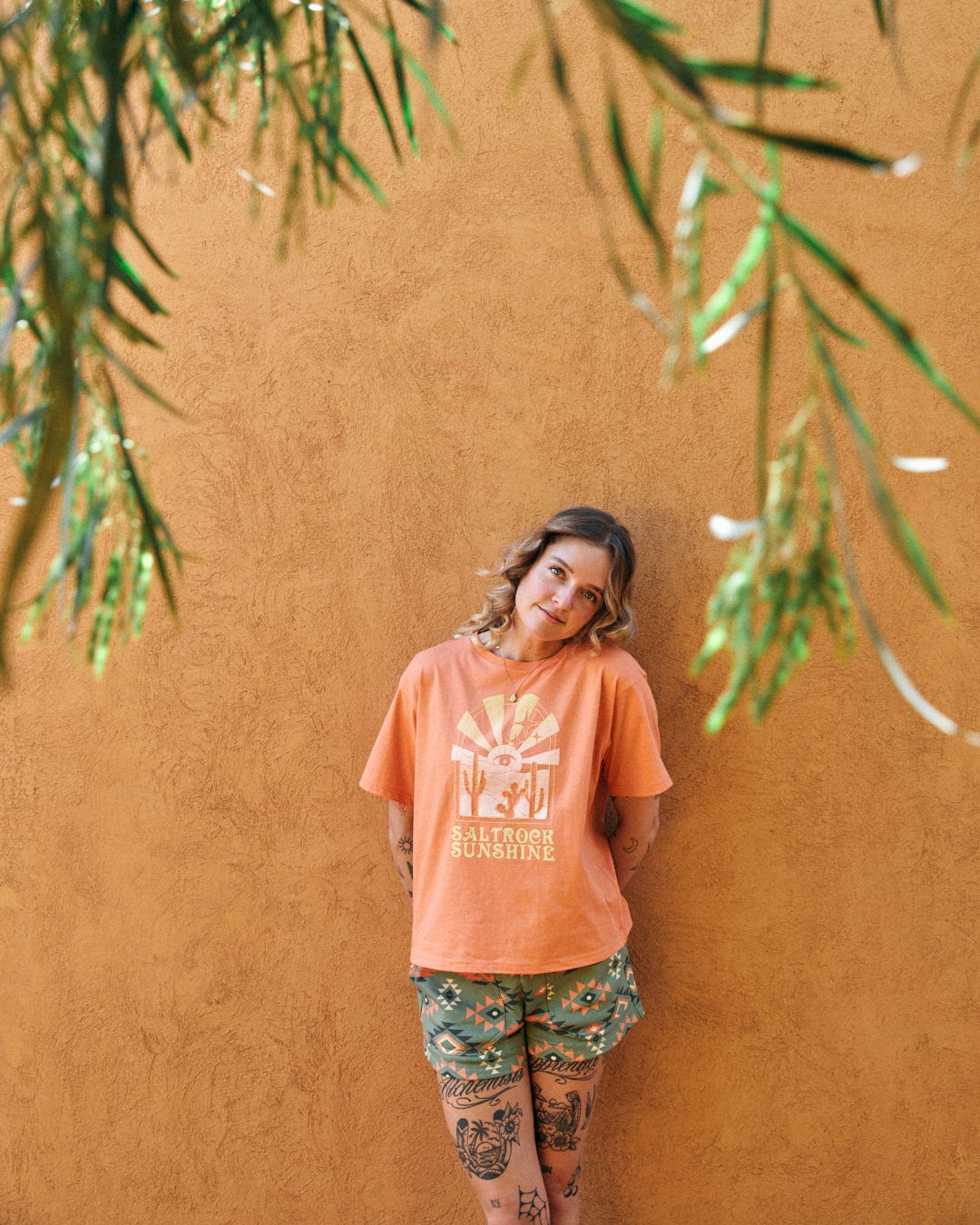 A person leans against an orange wall, wearing an orange t-shirt with Saltrock branding and patterned shorts. Featuring a boxy design with relaxed drop shoulders, the outfit blends seamlessly into the mystic desert sunset backdrop. Green foliage hangs in the foreground.
