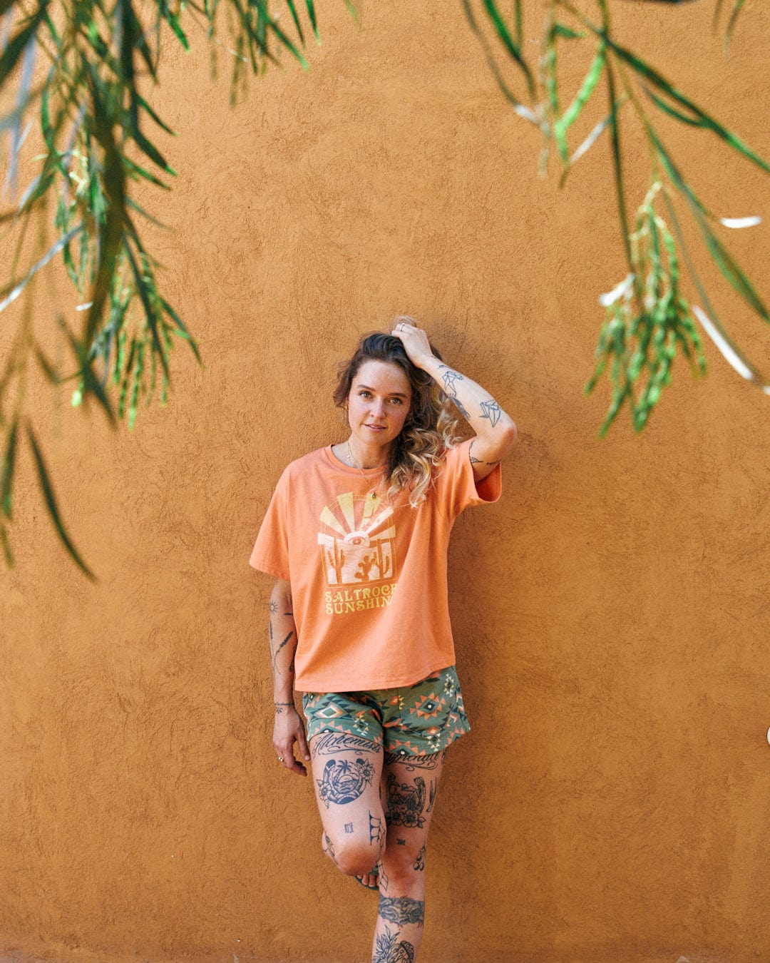 A person with tattoos, wearing an orange Saltrock Sunshine - Recycled Womens Cropped T-Shirt - Orange with Saltrock branding and patterned shorts, stands against an orange wall while touching their hair. Green leaves frame the top of the image.
