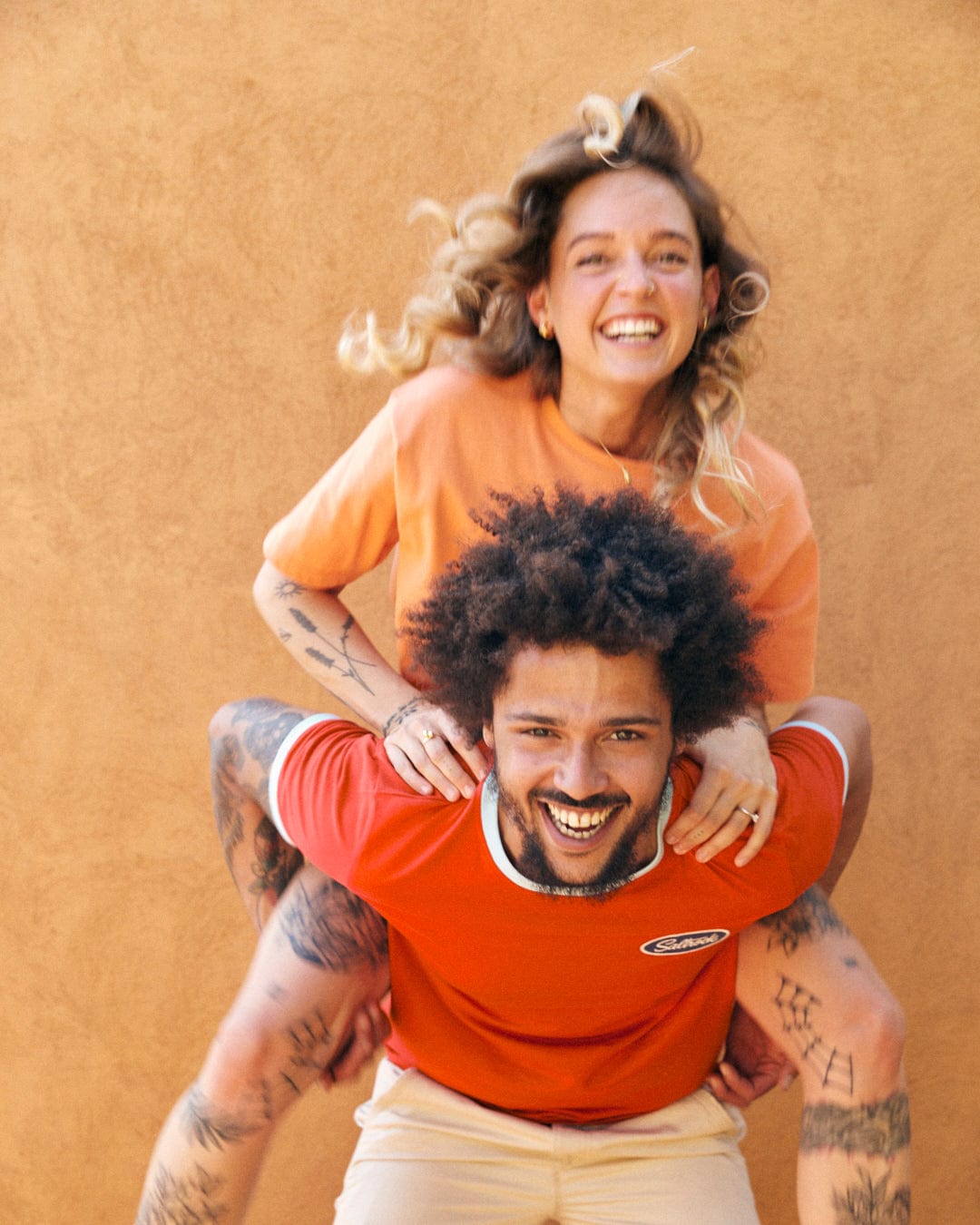 A smiling woman with blonde hair and an orange Saltrock Sunshine - Recycled Womens Cropped T-Shirt - Orange is piggybacking on a smiling man with curly hair and a red shirt, both showcasing their tattoos. The scene emits Saltrock branding vibes against the plain brown wall backdrop.