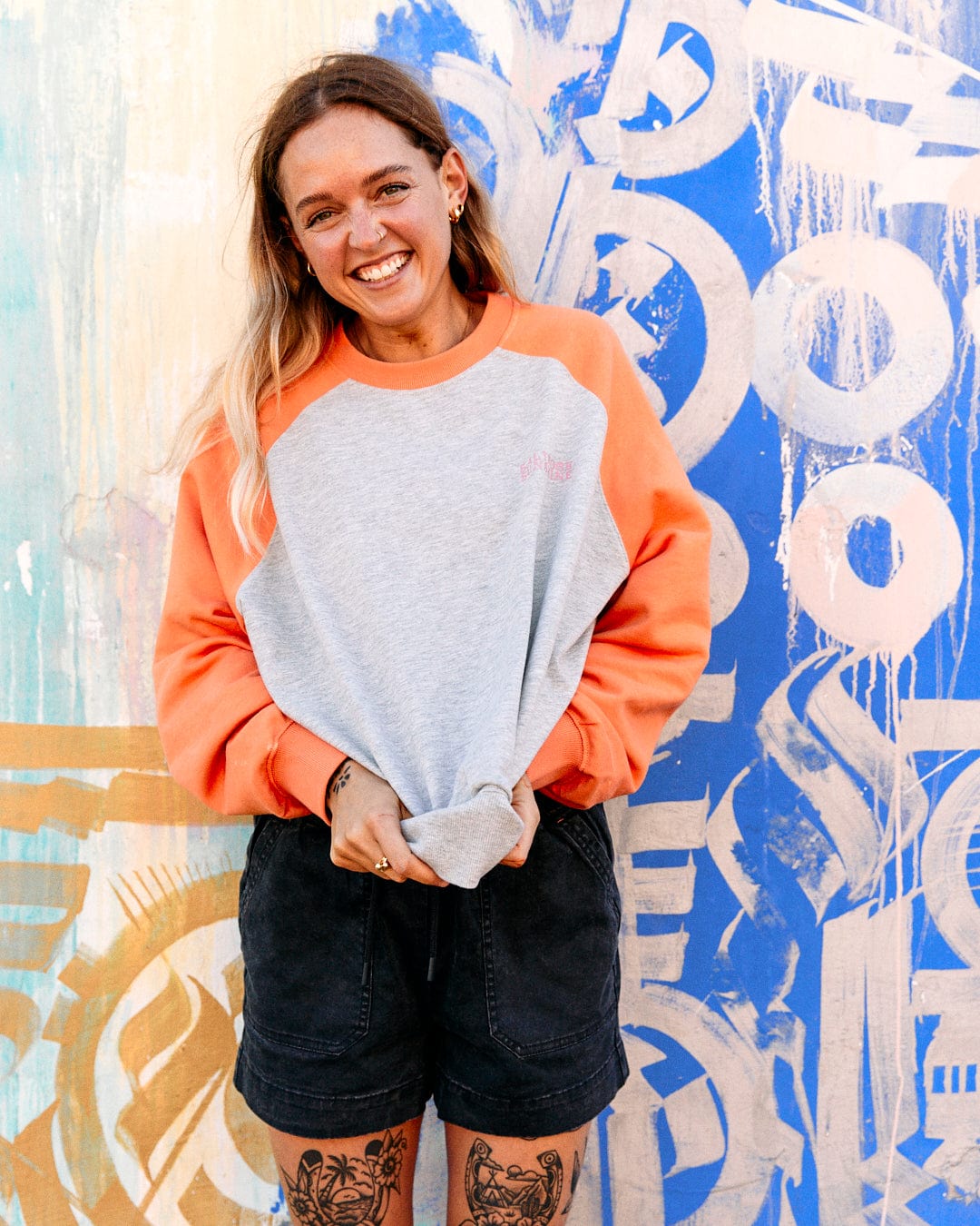 A cheerful young woman in a Saltrock Sunshine - Womens Raglan Sweat - Grey Marl and black shorts standing in front of a blue graffiti wall.