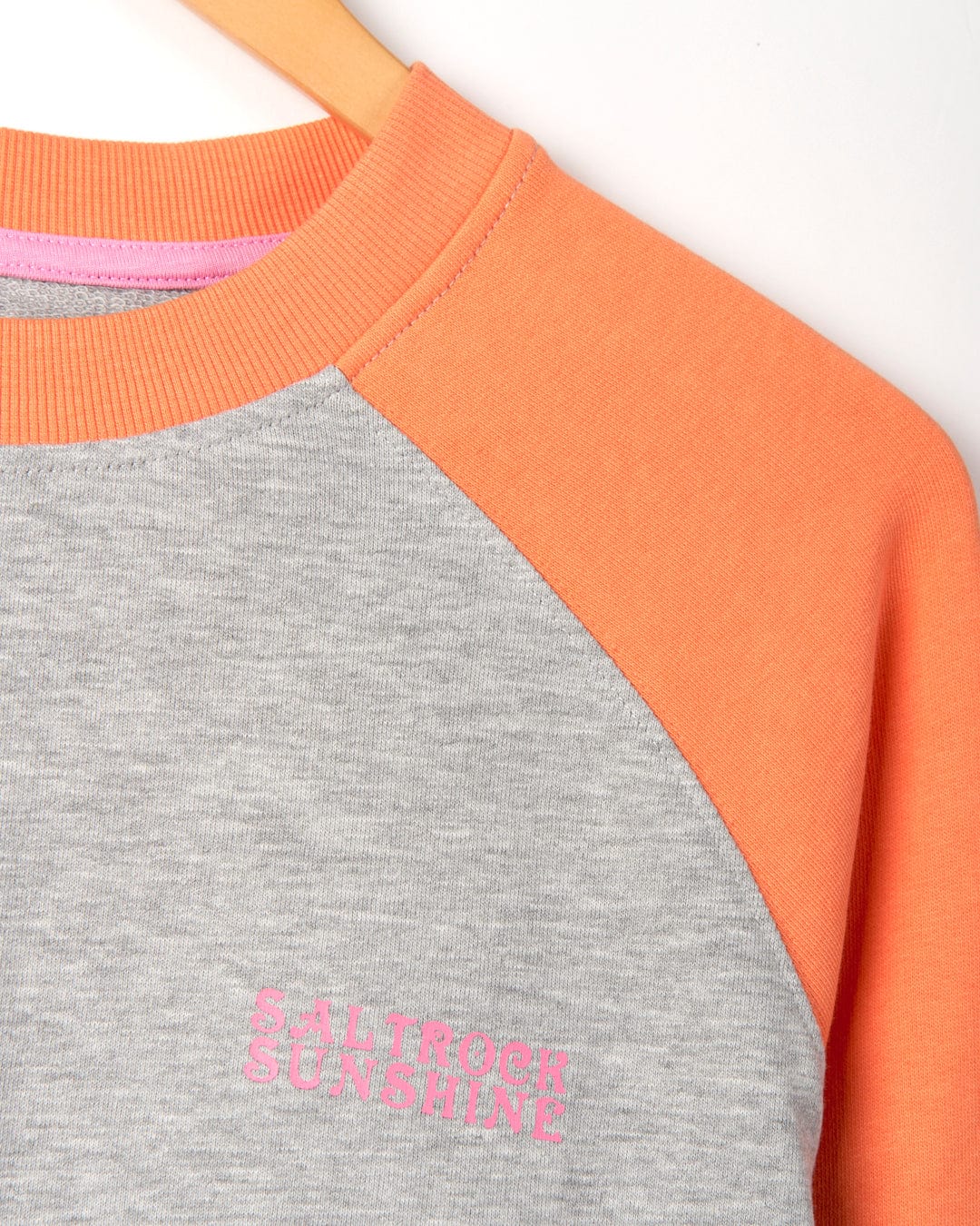 Close-up of a two-toned Saltrock Sunshine - Womens Raglan Sweat - Grey Marl sweatshirt, with gray on the left and orange on the right, hanging on a wooden hanger. The text "Saltrock Sunshine" is embroidered on the gray side, which features a marl effect fabric.