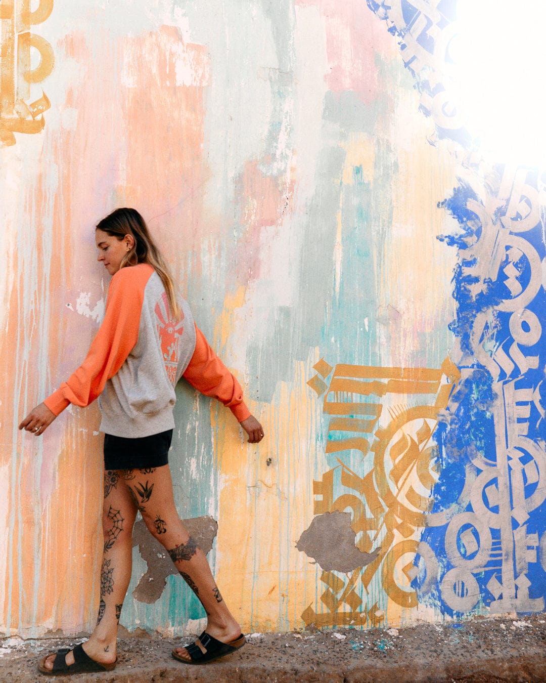 A woman walks past a colorful graffiti wall, wearing an orange Saltrock Sunshine - Womens Raglan Sweat - Grey Marl sweatshirt and black shorts, viewed in profile.