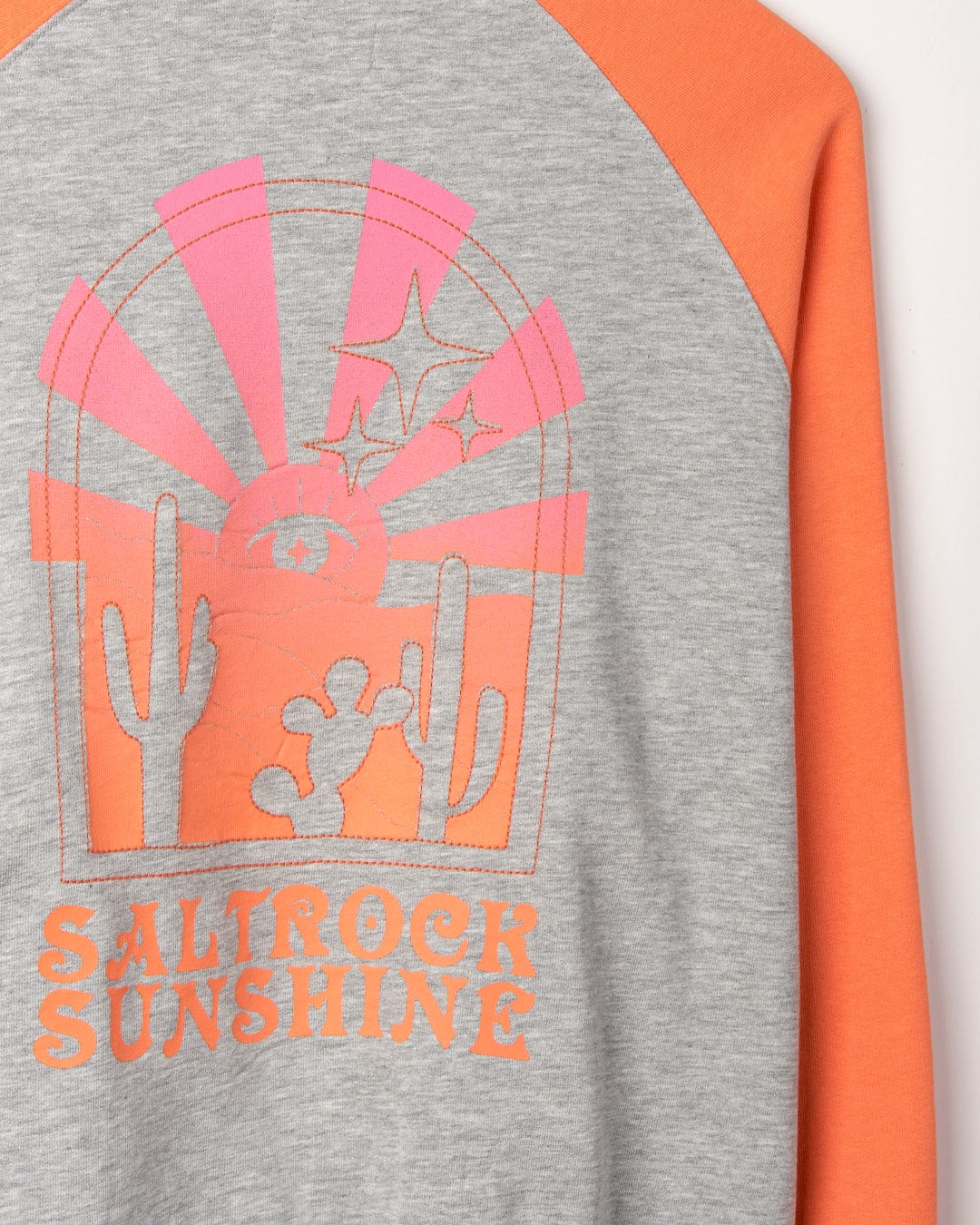 Close-up of a Saltrock Sunshine - Womens Raglan Sweat - Grey Marl fabric shirt with a graphic of a sunrise, cacti, and stars, and the text "Saltrock Sunshine" in orange.