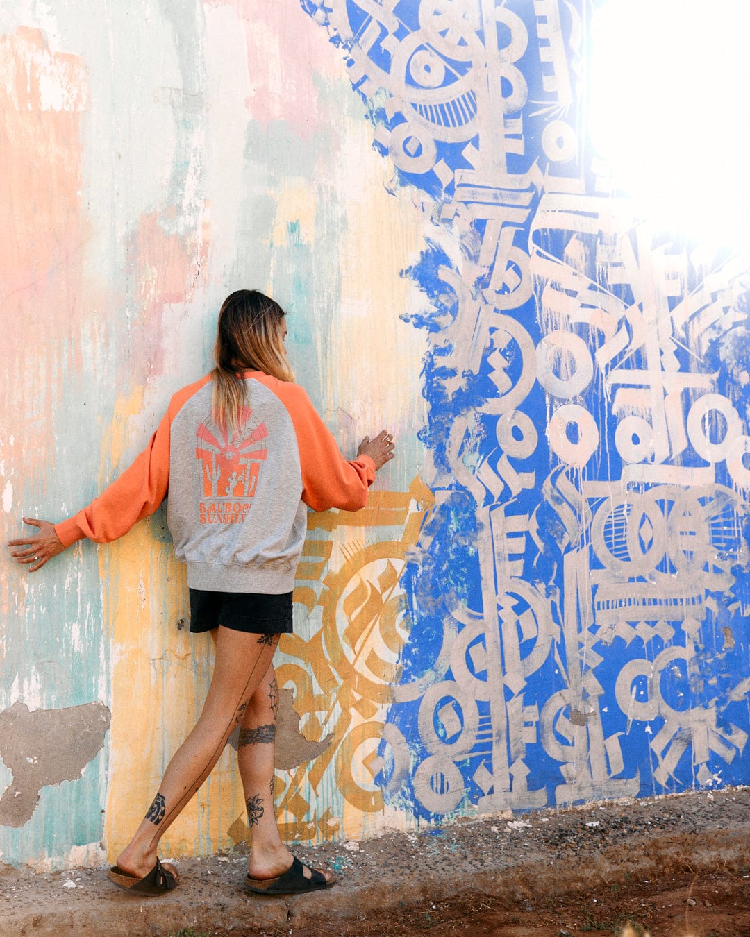 A woman in a Saltrock Sunshine - Womens Raglan Sweat - Grey Marl hoodie with raglan sleeves and black shorts touches a colorful mural wall featuring vibrant abstract designs.