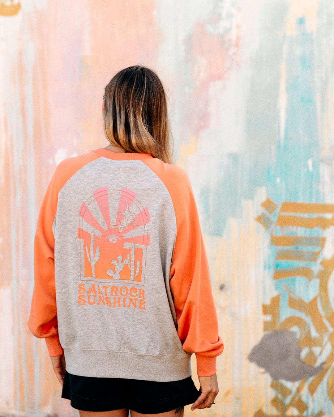Woman in a Saltrock Sunshine - Womens Raglan Sweat - Grey Marl with raglan sleeves and a graphic design on the back, standing in front of a colorful, abstract mural.