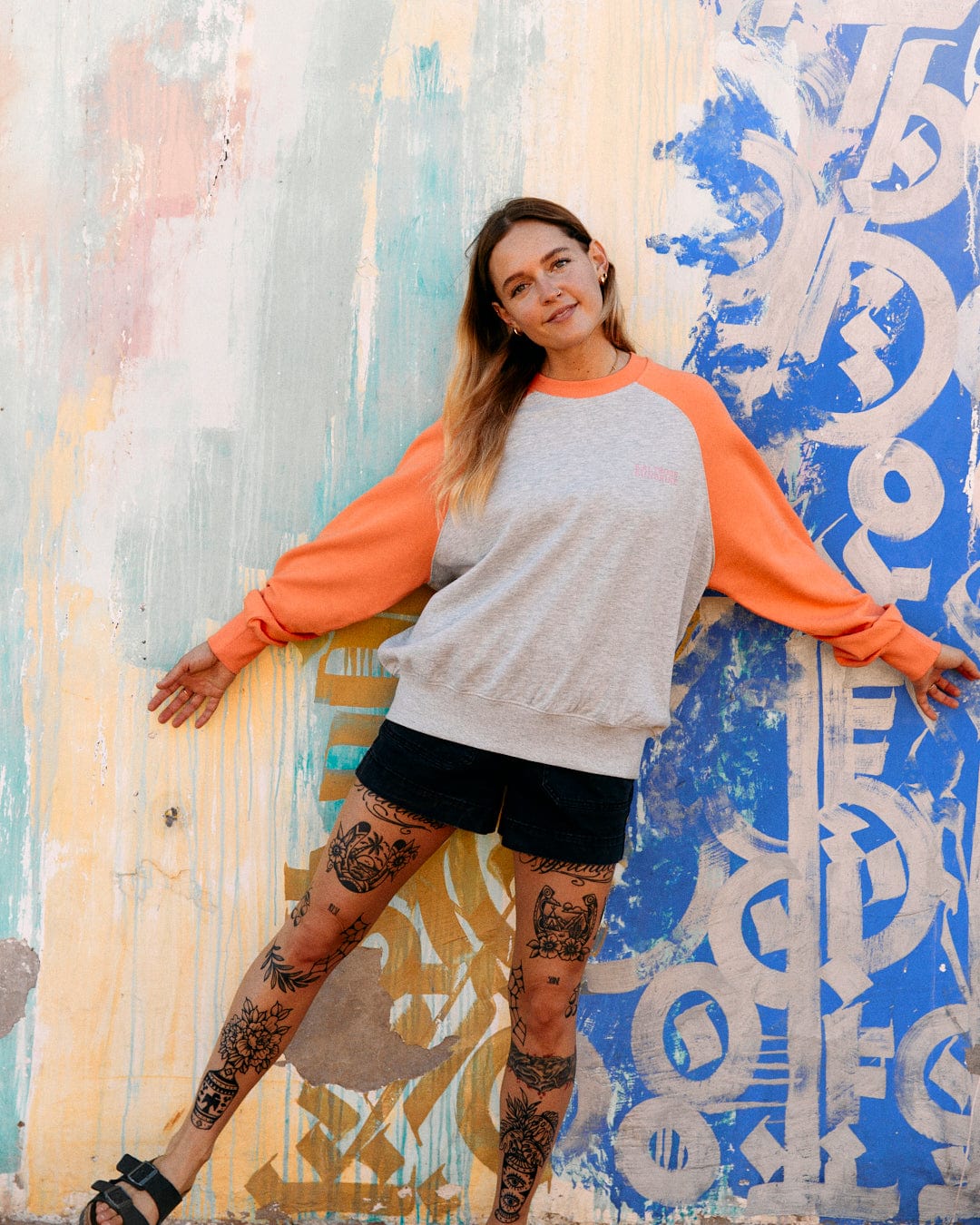 A woman with tattooed legs stands against a graffiti-covered wall, wearing a colorful Saltrock Sunshine sweatshirt with raglan sleeves and black shorts.