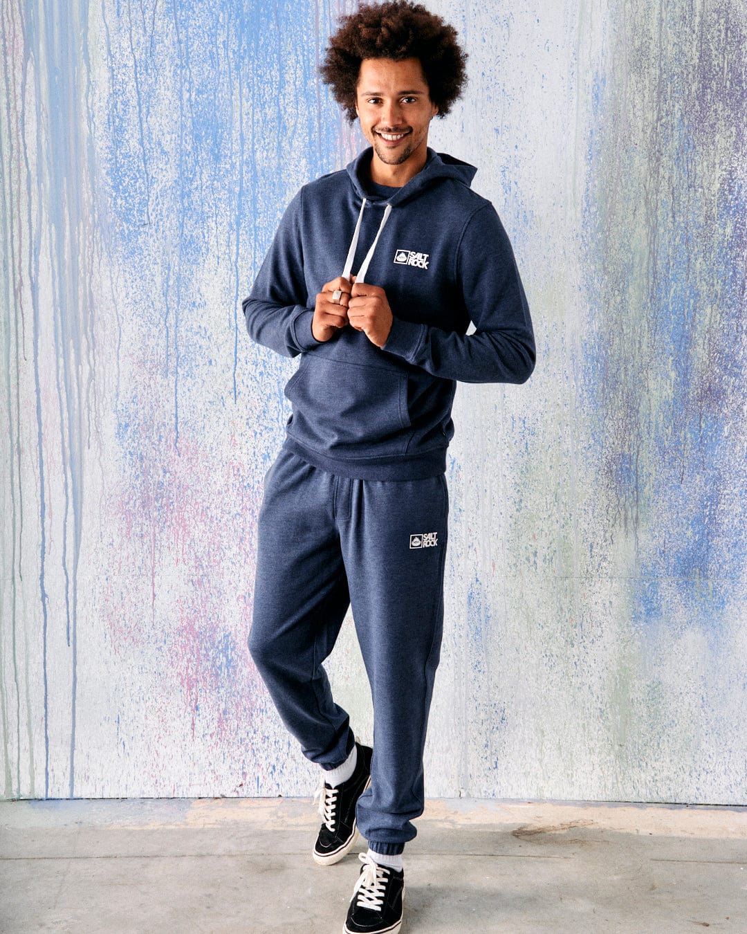 A man with curly hair smiles while wearing a Saltrock dark blue hoodie with a super soft lining and matching Saltrock Original Men's Joggers featuring an elasticated waist, standing in front of a colorful, textured wall.