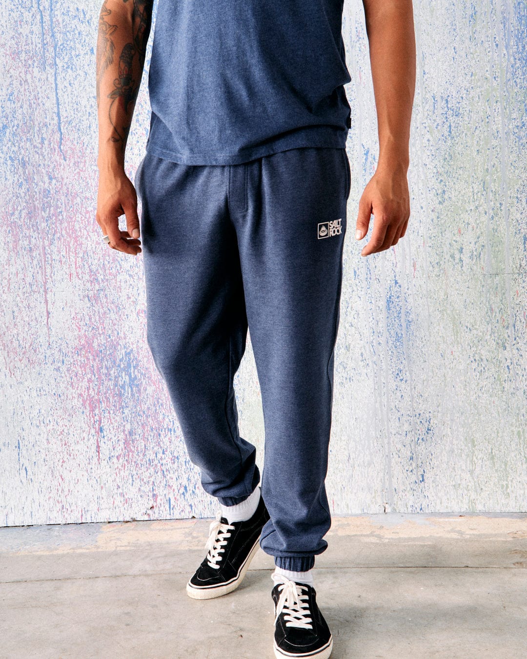 A person in a blue short-sleeve shirt and Saltrock Original Men's Joggers in Dark Blue with an elasticated waist stands on a concrete floor against a multicolored wall. The person wears black sneakers, and their right arm has visible tattoos.