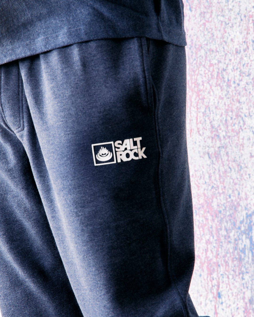 Close-up view of a person wearing the Saltrock Original - Mens Joggers in Dark Blue, featuring the "Saltrock" logo printed on the leg. Made from soft jersey material with an elasticated waist, these joggers offer comfort and style. The background features a textured, multicolored surface.