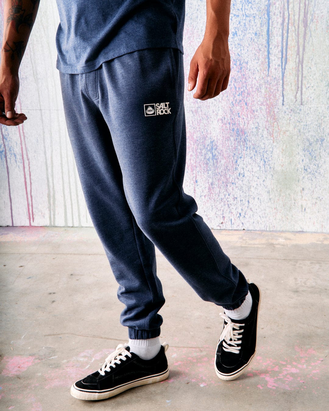 A person in a blue t-shirt and Saltrock Original Mens Joggers in Dark Blue, featuring an elasticated waist and a white "Saltrock" logo, stands on a concrete floor near a wall with colorful paint splatters. They are wearing black and white sneakers. The super soft lining of the joggers gives them a comfortable look.