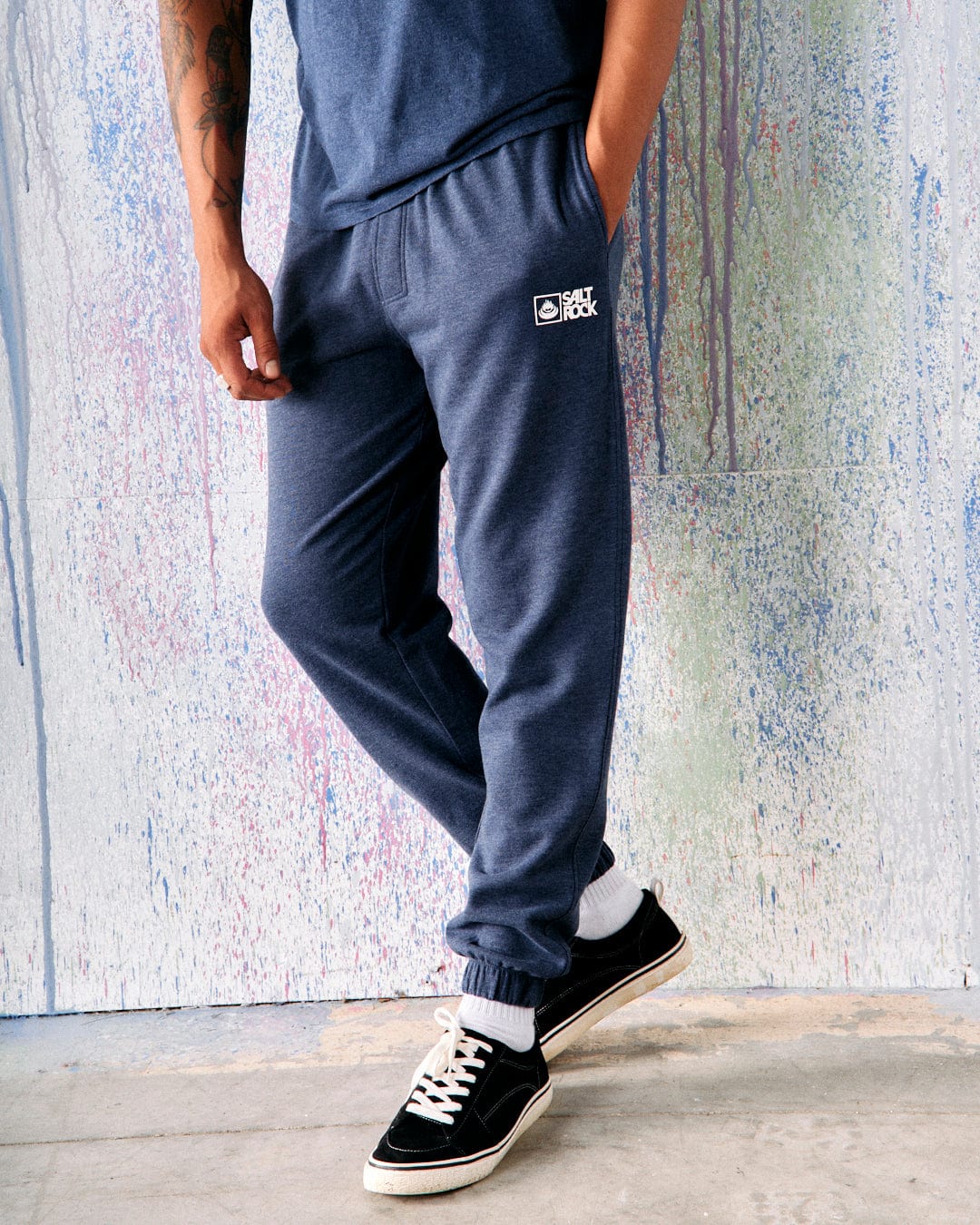 Person wearing Saltrock Original - Mens Joggers in dark blue, made of soft jersey material with their hands in pockets, standing in front of a multicolored splattered wall. Only lower body and part of the torso visible.