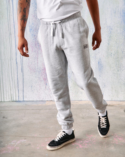 Person wearing Saltrock Original - Mens Joggers in grey made of soft jersey material, a white T-shirt, and black sneakers stands with one leg forward. The background features a painted, abstract design.