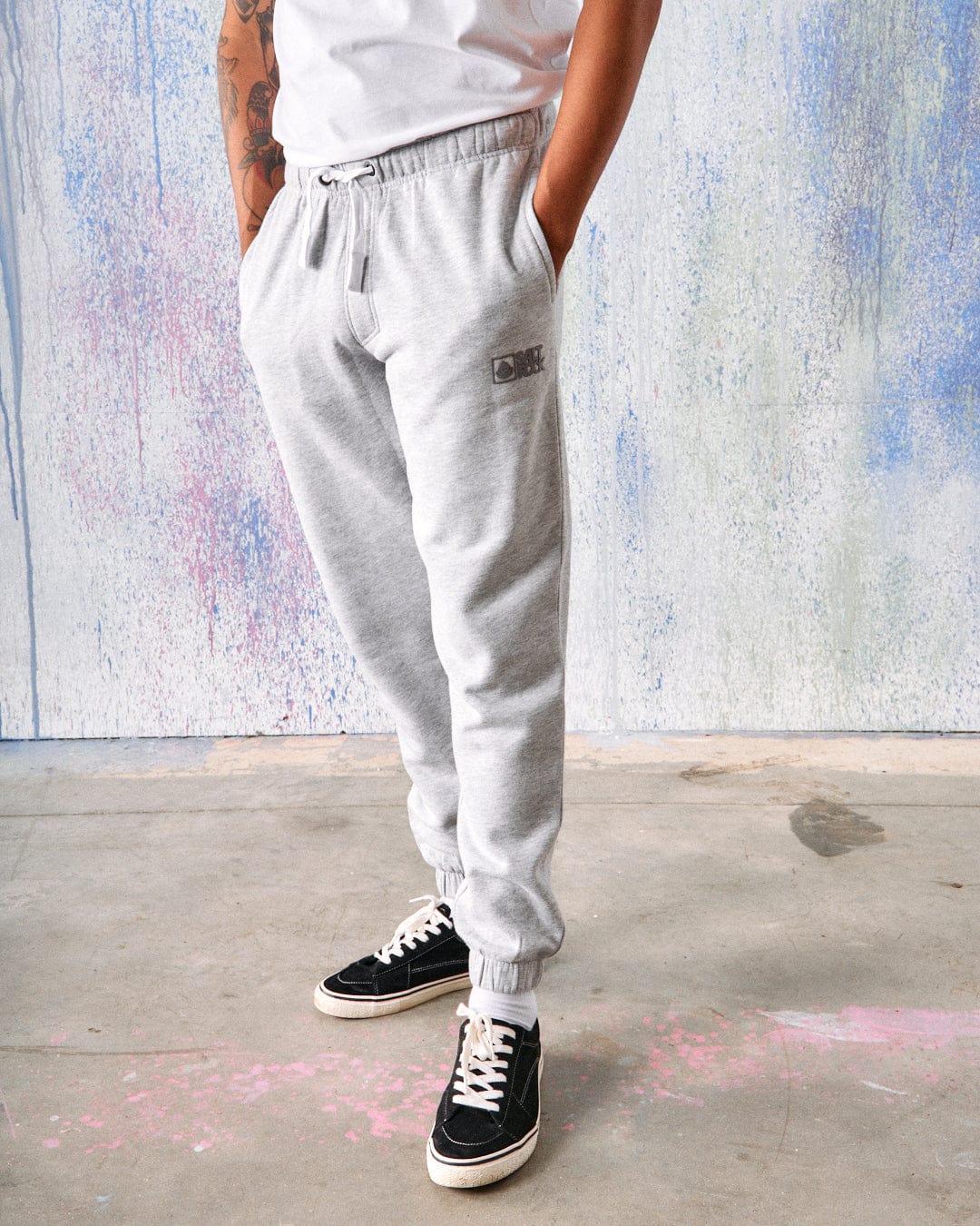 A person wearing Saltrock Original - Mens Joggers in grey, a white shirt, and black sneakers stands on a concrete floor with a multicolored wall in the background. Their hands are in their pockets.