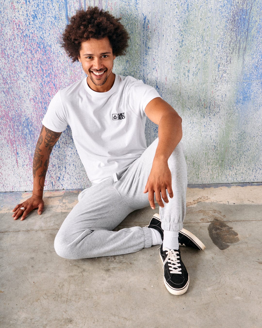 A man with curly hair and a tattoo sits on the floor against a colorful, speckled wall. Smiling at the camera, he wears a white t-shirt, Saltrock Original Men's Joggers in grey with an elasticated waist, and black sneakers.