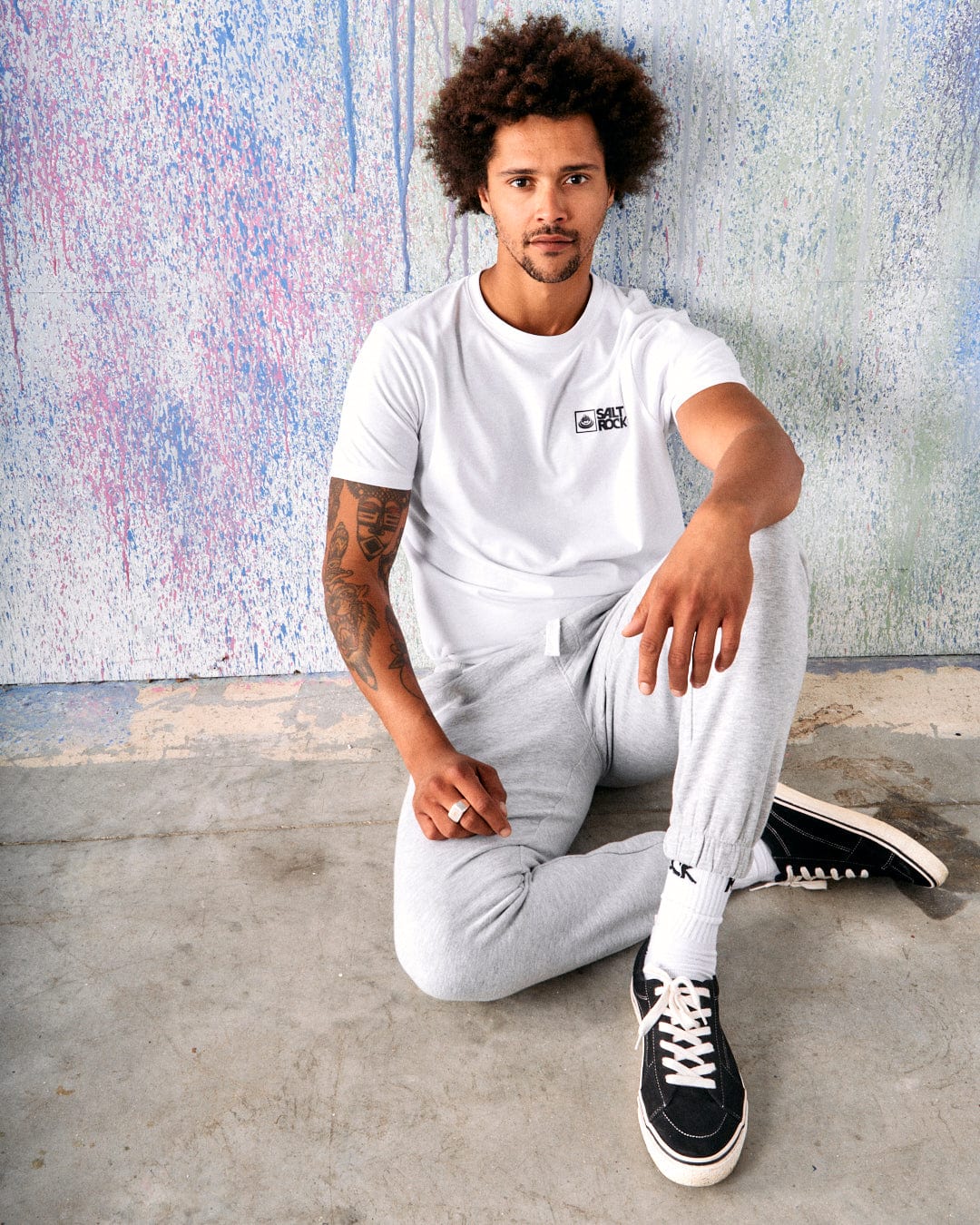 A man with curly hair and a beard sits on the floor against a colorful wall. He is wearing a white T-shirt, Saltrock Original - Mens Joggers in grey, and black sneakers. He has a tattoo on his left arm.