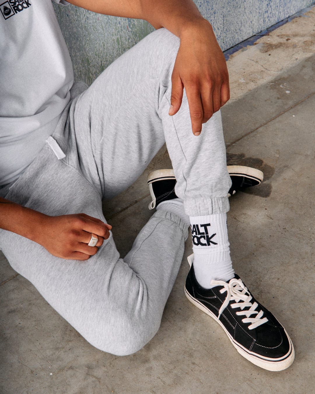 A person wearing the Saltrock Original Men's Joggers in grey, made of soft jersey material, and white socks with "ALT ROCK" printed on them, sits on the ground with one hand resting on their knee and the other on the floor. Their black sneakers add a contrasting touch.