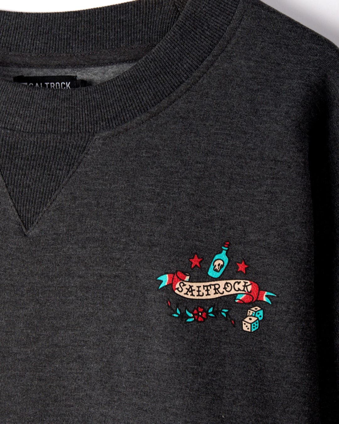 A close-up of a dark grey **Sea Siren - Mens Sweat - Dark Grey** with a colorful **Saltrock** logo featuring a bottle, dice, and flowers embroidered on the chest, showcasing an oversized fit.