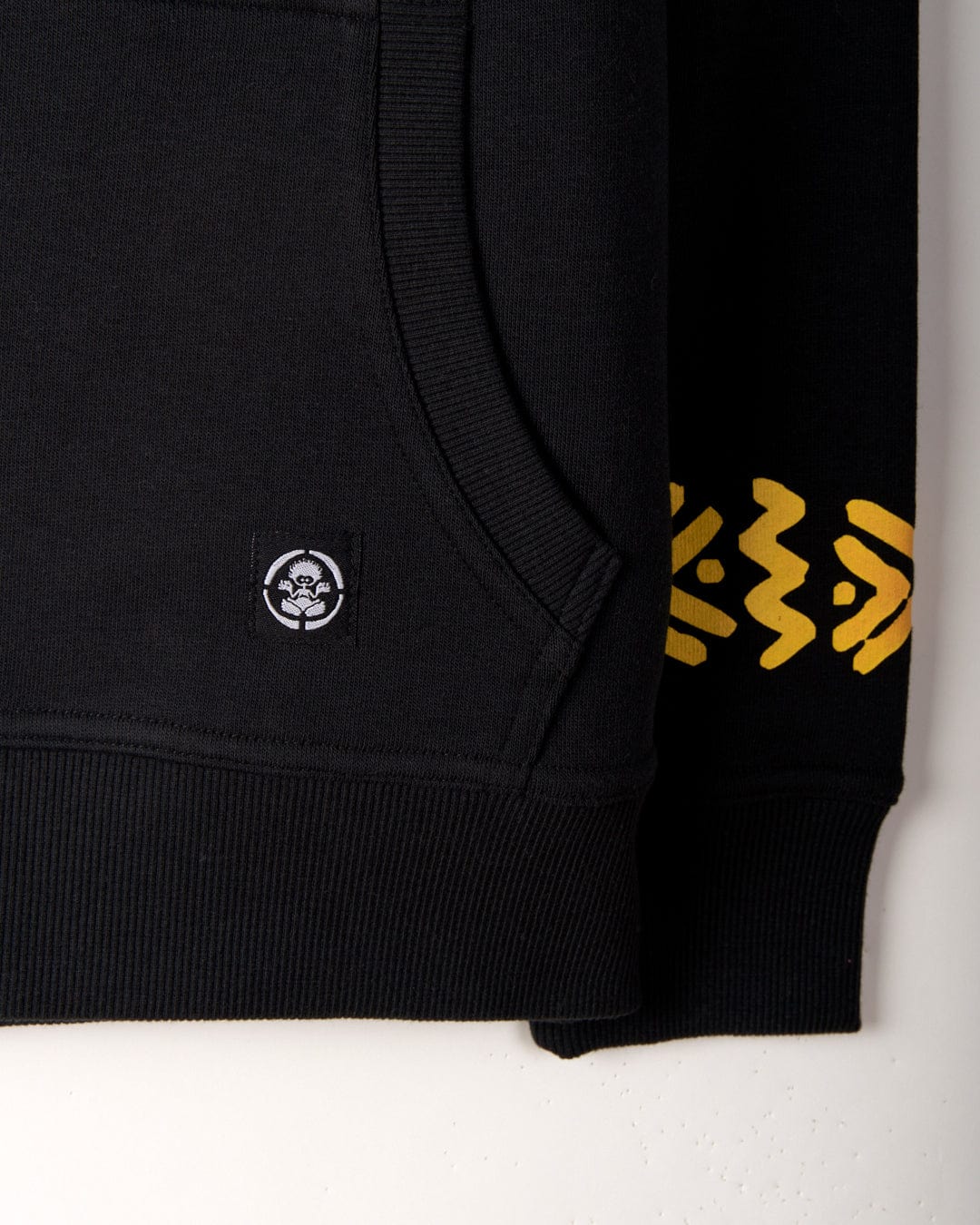 Close-up of a black hoodie featuring Saltrock branding with a small circular logo on the kangaroo pocket and yellow abstract patterns on the sleeve. The product is the See No Skulls - Kids Recycled Pop Hoodie - Black by Saltrock.