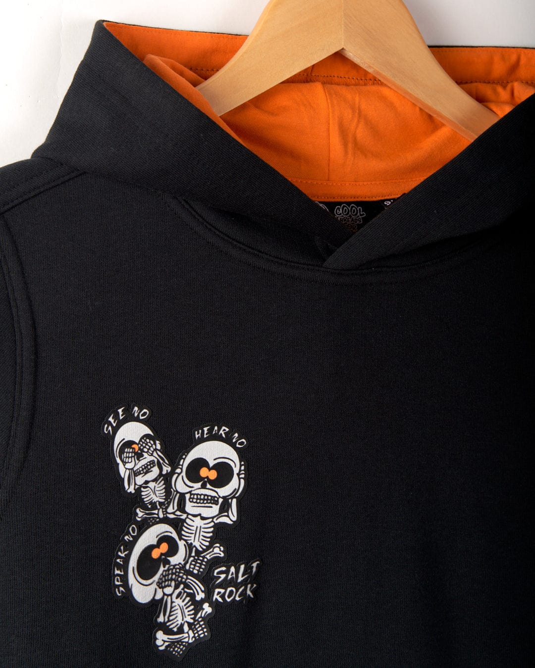 A black hoodie with an orange inner lining displayed on a wooden hanger. It features three skeleton illustrations and text that reads "SPEAK NO, HEAR NO, SEE NO," along with distinctive Saltrock branding. The design includes a convenient kangaroo pocket for added functionality. Known as the See No Skulls - Kids Recycled Pop Hoodie - Black by Saltrock, this product offers both style and practicality.