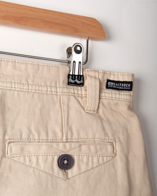 Close-up of beige cotton chinos on a wooden hanger, focusing on the back pocket and brand label "Saltrock" on the waistband. These Sennen - Mens Chino Short - Cream from Saltrock are perfect for everyday wear.