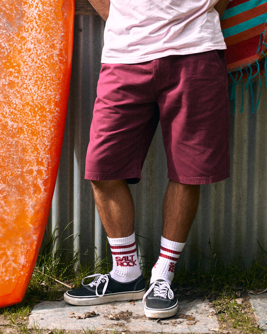 A person wearing a relaxed fit white T-shirt and *Saltrock* burgundy chino shorts stands with their hands behind their back. They complete the look with white "Salt Rock" socks, black sneakers, and an orange surfboard propped nearby.