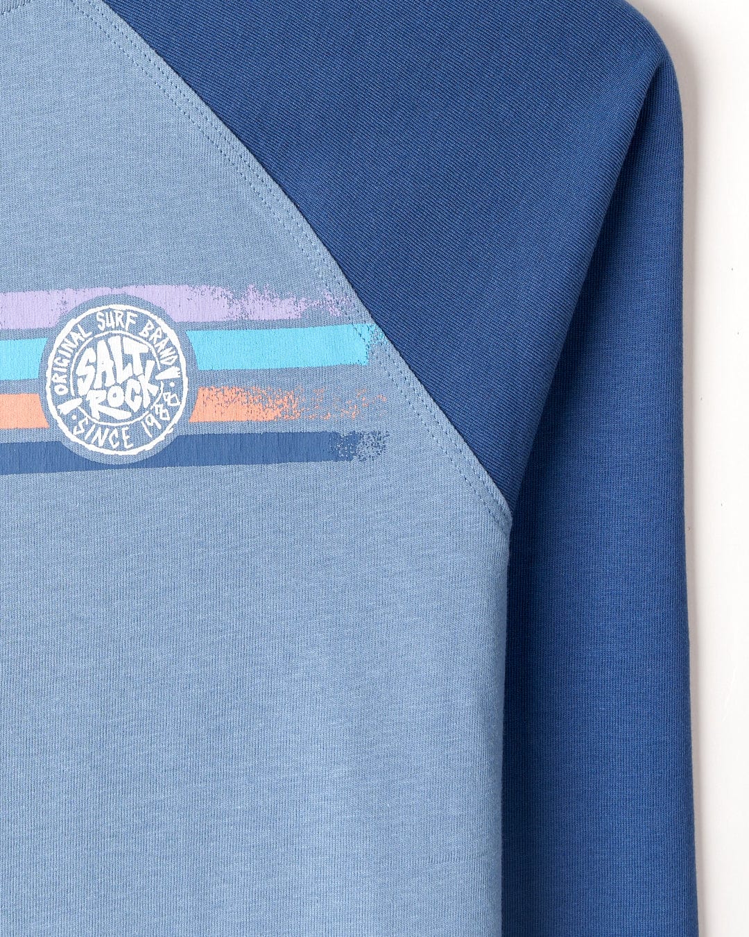 Close-up of a blue Spray Stripe - Kids Raglan Long Sleeve T-Shirt with Saltrock branding featuring text "original silk road salt rock 1986" in white, pink, and light blue.