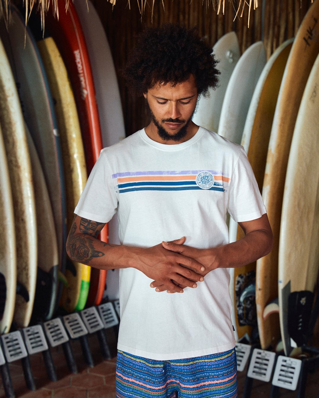 A man stands with his hands clasped in front of a rack of surfboards, wearing the Saltrock Spray Stripe - Mens Short Sleeve T-Shirt - White and patterned blue shorts, proudly displaying the Saltrock branding.