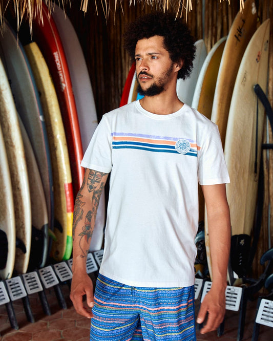 A man with a beard and tattoos stands indoors, wearing a Spray Stripe - Mens Short Sleeve T-Shirt - White and blue shorts. Behind him are various surfboards lined up against the wall, while his 100% cotton attire proudly displays Saltrock branding.