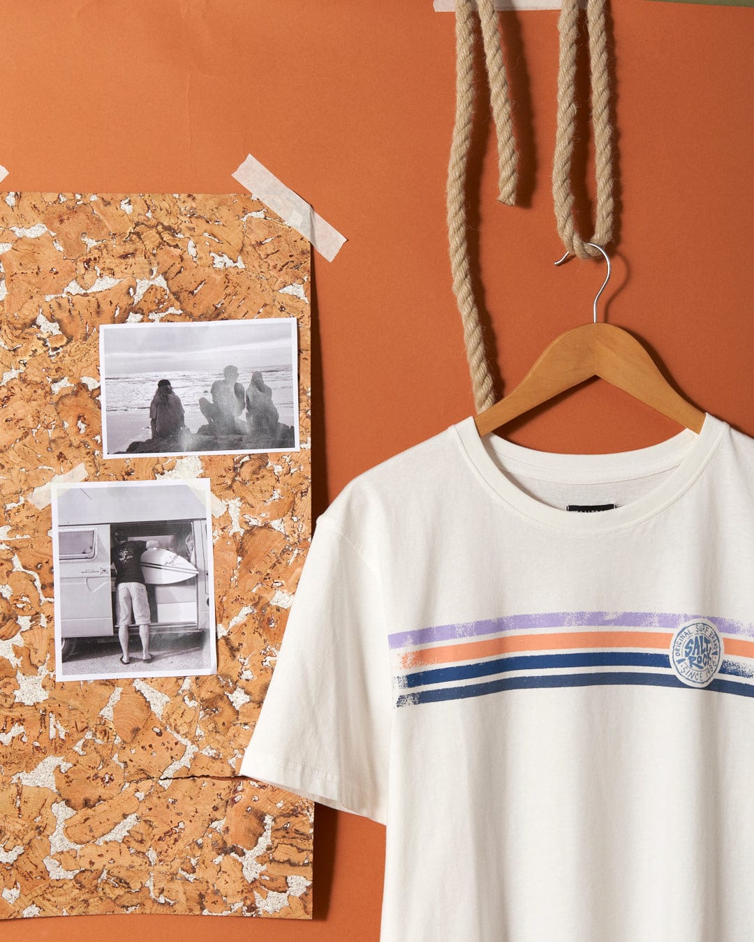 A white Spray Stripe - Mens Short Sleeve T-Shirt - White with colorful stripes and Saltrock branding on a hanger is displayed beside a corkboard with two black-and-white photographs, all against an orange background with a coiled rope hanging above.
