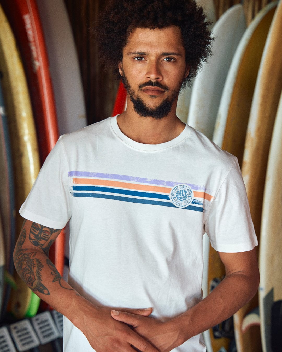 A man with a beard and curly hair stands in front of multiple surfboards, wearing the Spray Stripe - Mens Short Sleeve T-Shirt - White with Saltrock branding.