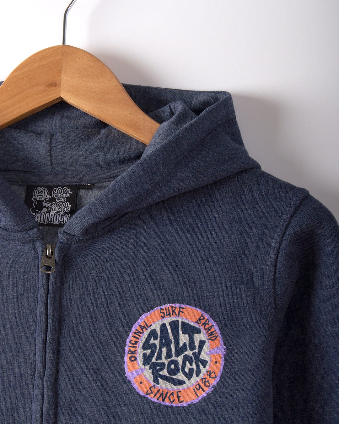 Navy blue Saltrock hoodie on a wooden hanger, showcasing the "Original Surf Brand Since 1988" text on the chest. The SR Original - Kids Zip Hoodie is crafted from soft cotton, and its hood provides an extra layer of comfort for those breezy beach days.