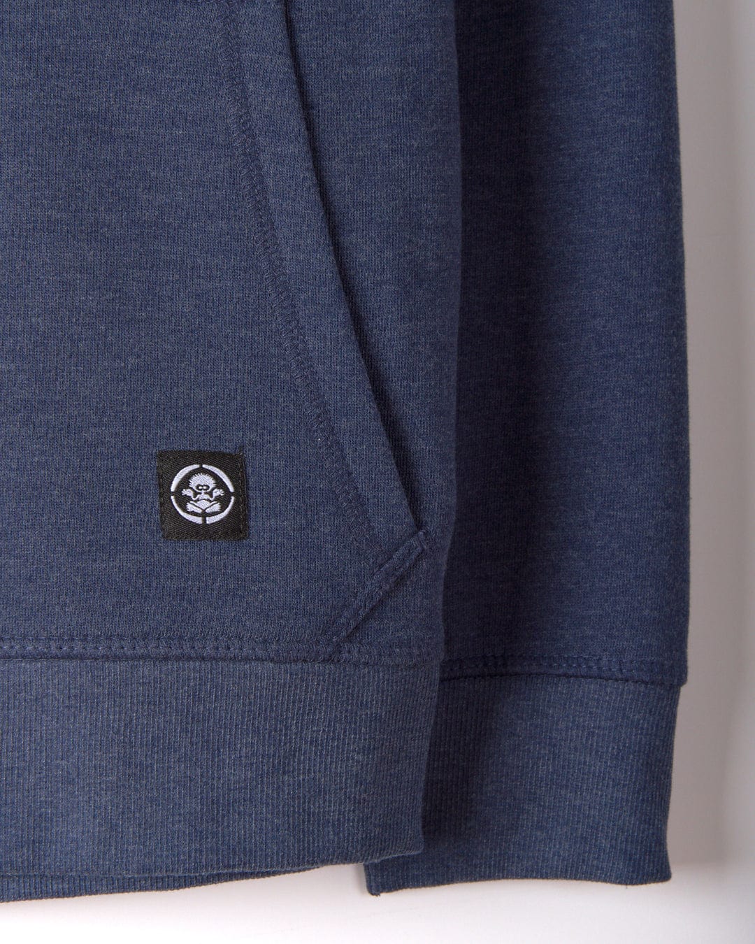 Close-up of a blue SR Original Kids Zip Hoodie by Saltrock, showcasing a black and white logo patch with a stylized face near the pocket.