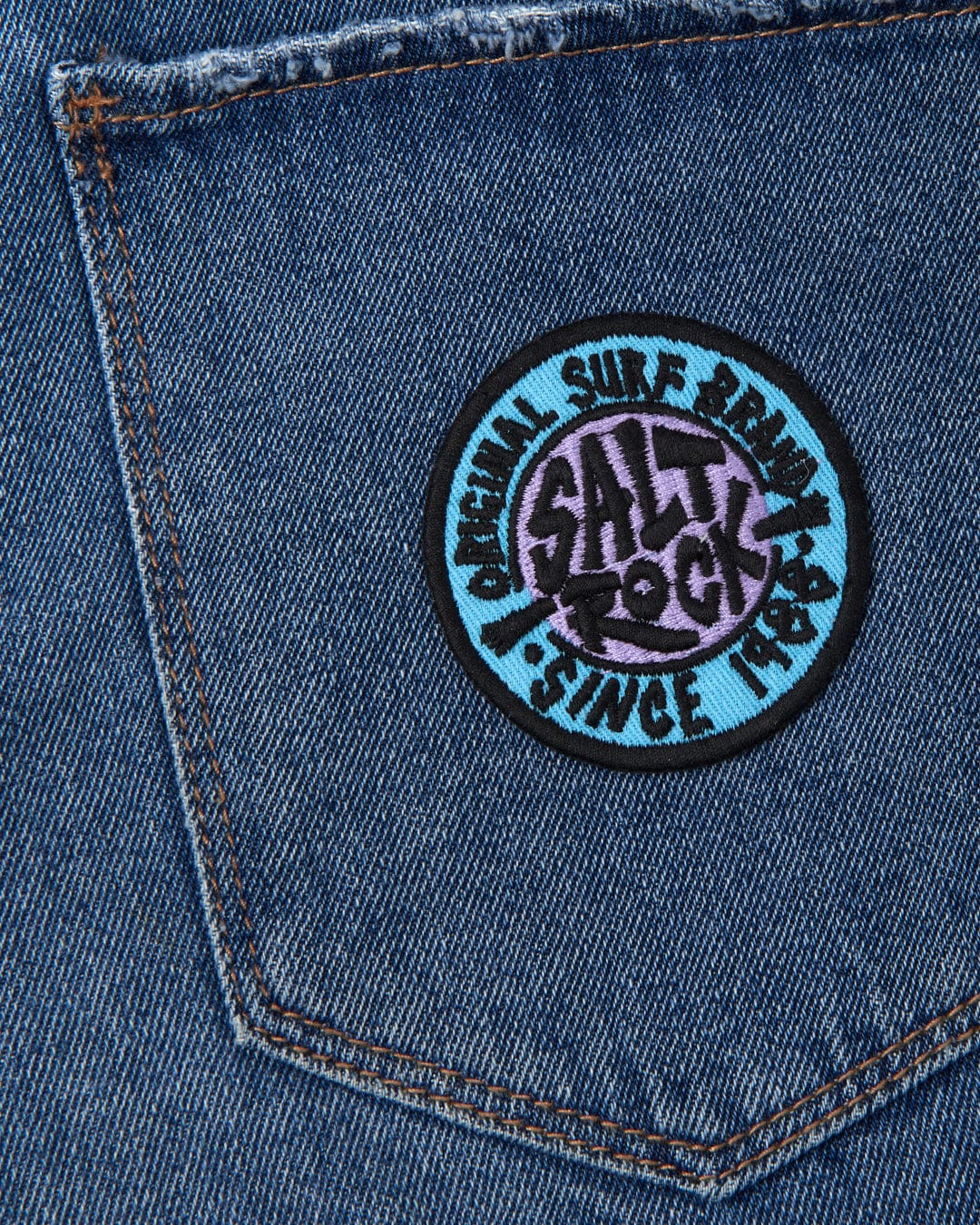 A close-up of a denim fabric with a circular Saltrock branding iron-on patch featuring the SR Original - Patches - Blue design.