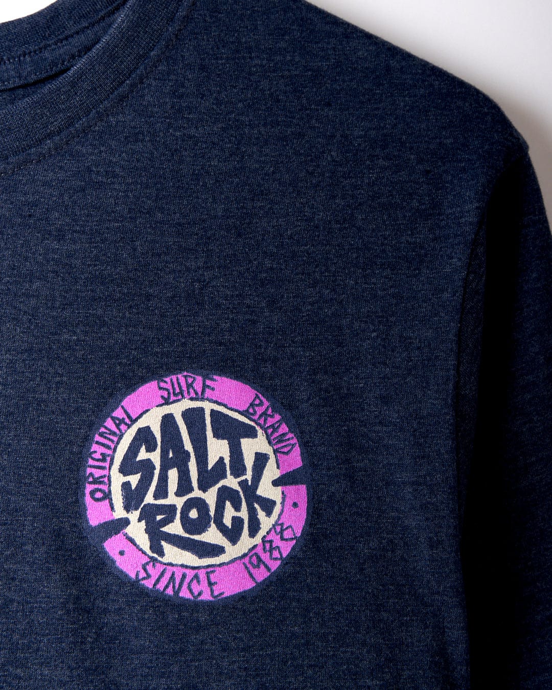 Close-up of the SR Original - Mens T-Shirt - Blue from Saltrock featuring a circular logo with "Original Surf Brand Since 1988" text in purple and white. Made with 60% cotton, this Saltrock t-shirt offers a comfortable crew fit perfect for any casual outing.