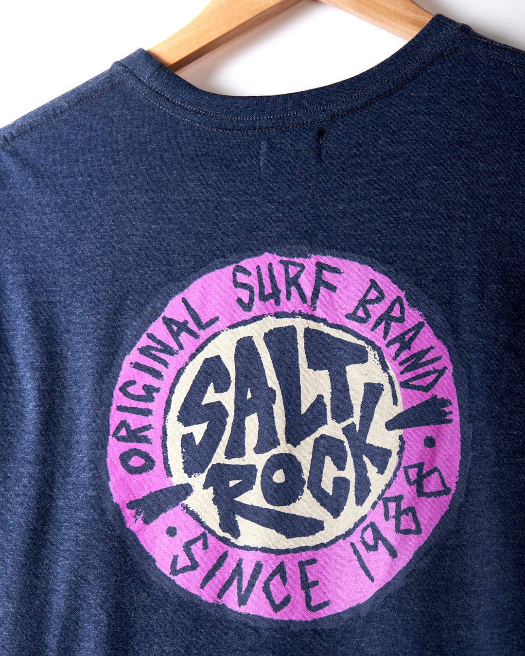 Close-up of the SR Original men's t-shirt by Saltrock, showcasing its navy blue crew fit design. The back features a circular graphic with the text "Salt Rock, SR Original Surf Brand, Since 1988" in pink and white. Made from 60% cotton, the shirt is hanging on a wooden hanger.