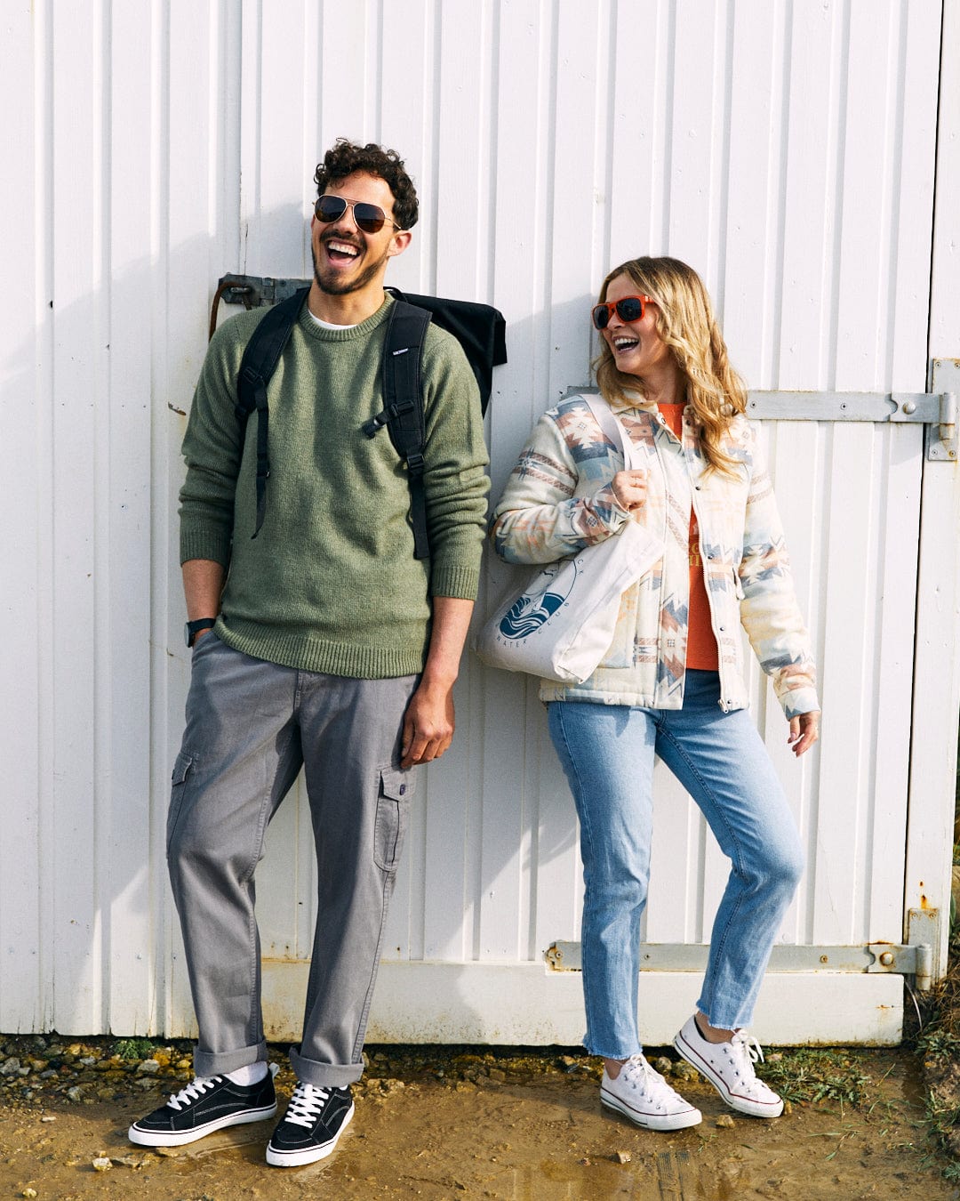 Two people are standing and laughing in front of a white wooden wall, both wearing sunglasses. The person on the left has a backpack, while the person on the right is holding a super soft, reusable bag and wearing a Saltrock Bowen - Mens Long Sleeve Crew Knit - Green.