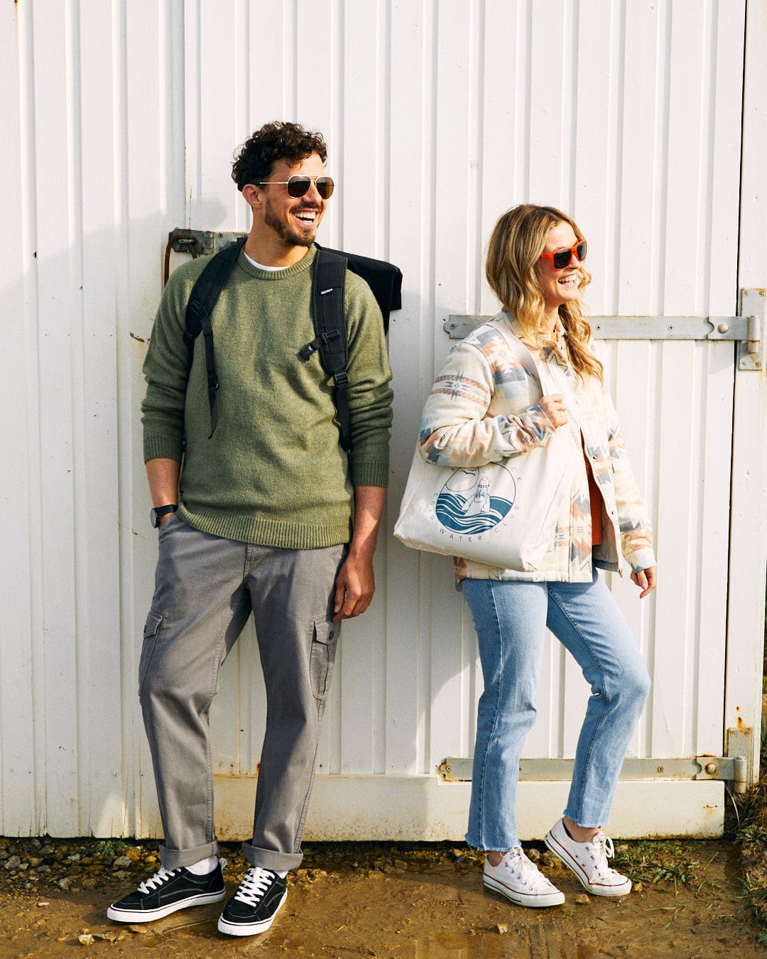 A man and a woman stand outside against a white wall. The man, in his machine washable Saltrock Bowen - Mens Long Sleeve Crew Knit - Green, gray pants, and sunglasses, carries a backpack. The woman sports sunglasses too, along with her super soft patterned jacket, blue jeans, and a tote bag.