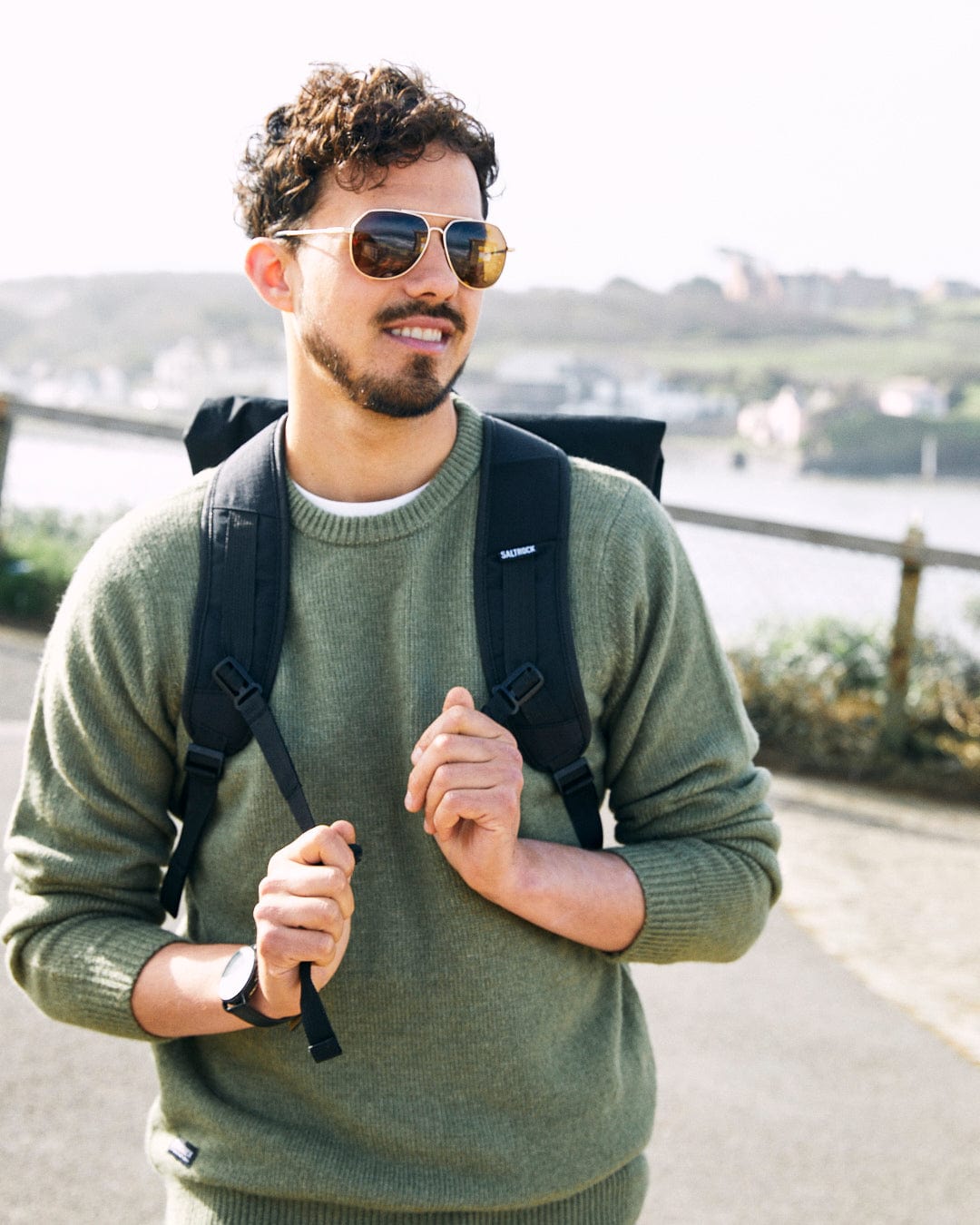 A man with curly hair and sunglasses wears a green, super soft Saltrock Bowen - Mens Long Sleeve Crew Knit and backpack. He is standing outdoors on a walkway with a view of water and hills in the background.