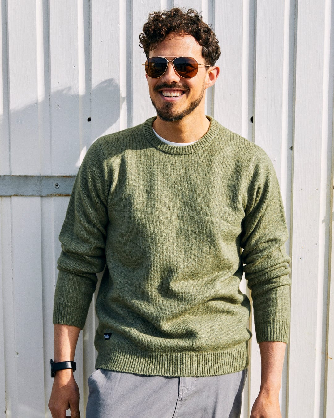 A man with curly hair and sunglasses smiles while wearing a green Saltrock Bowen - Mens Long Sleeve Crew Knit and standing in front of a white wooden wall.