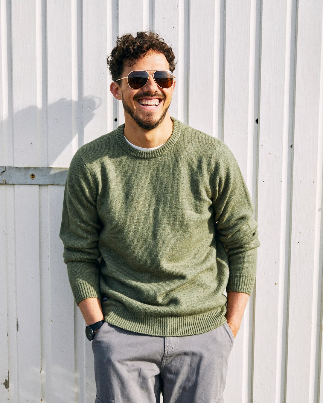 A man with curly hair and sunglasses stands against a white wooden wall, smiling. He wears a super soft, green Saltrock Bowen - Mens Long Sleeve Crew Knit and gray pants, with his hands in his pockets.