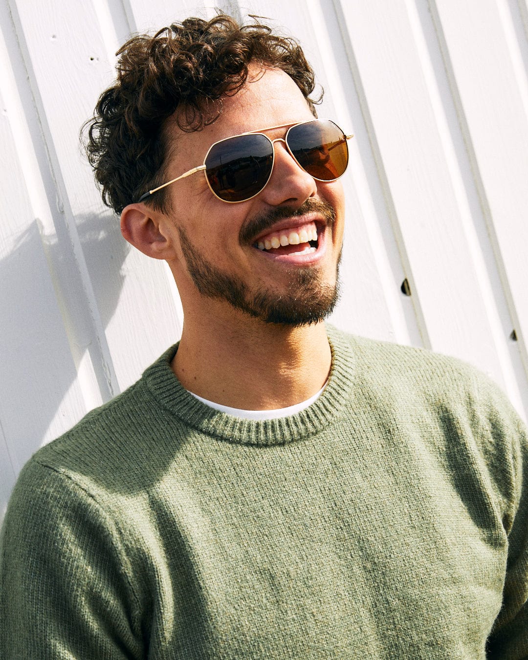 A man with curly hair and a beard, wearing sunglasses and a green, super soft Saltrock Bowen - Mens Long Sleeve Crew Knit - Green sweater, smiles while standing against a white wall.