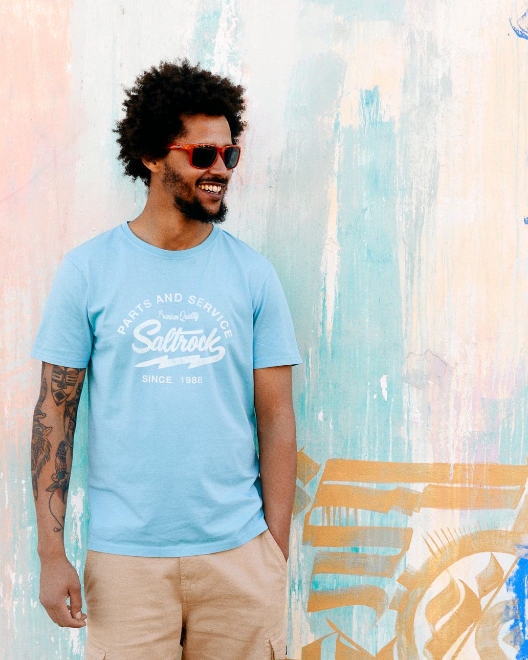 A man with curly hair and sunglasses, smiling, wearing a Last Stop Motel - Mens T-Shirt - Light Blue with Saltrock branding and beige shorts, stands against a graffiti-covered wall.