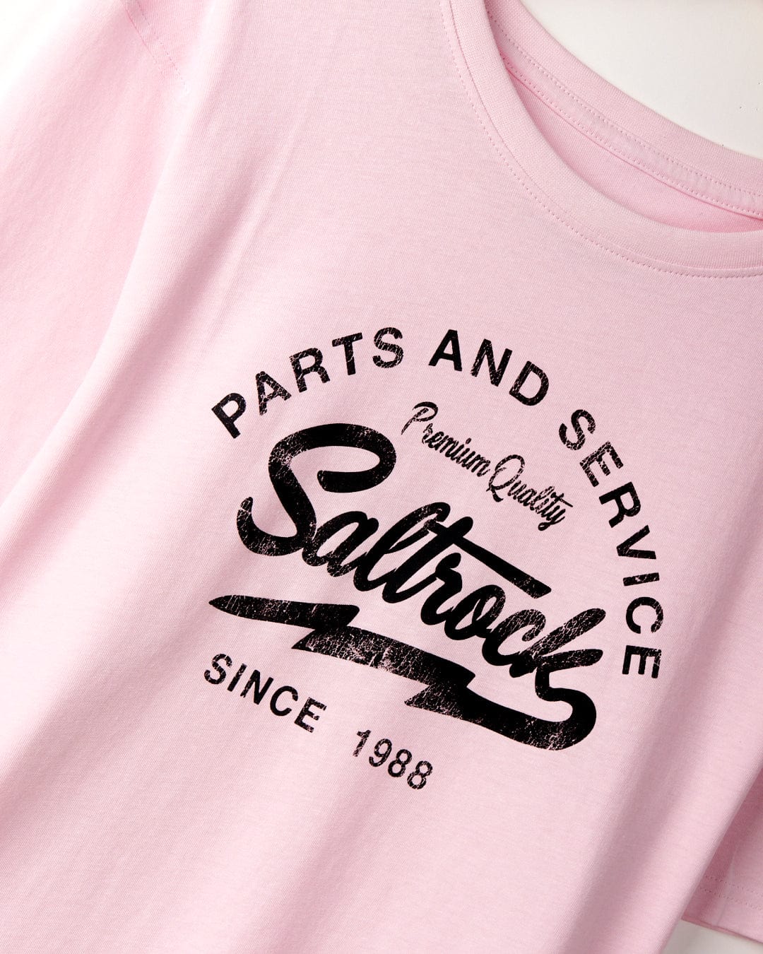 Close-up of a pink T-shirt showcasing Saltrock branding with "Parts and Service," "Saltrock," "Premium Quality," and "Since 1988" printed in black. Made from 100% cotton for ultimate comfort. This is the Strike Logo - Mens T-Shirt - Pink by Saltrock.