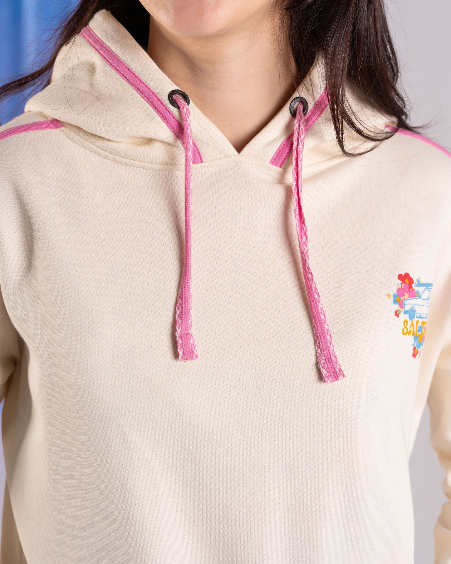 A person is wearing a Saltrock Highway women's pop hoodie made of soft cotton, featuring a cream color with pink drawstrings and a small colorful chest graphic.