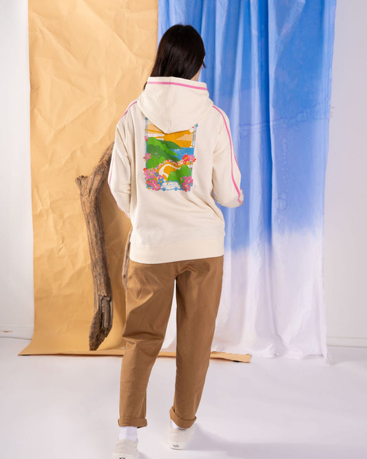 A person is standing with their back turned, dressed in the Saltrock Highway Women's Pop Hoodie in cream, featuring a colorful nature-themed design and a kangaroo pocket, paired with brown pants and sneakers. The textured beige and blue backdrop highlights hints of contrast pink detail among subtle graphics.