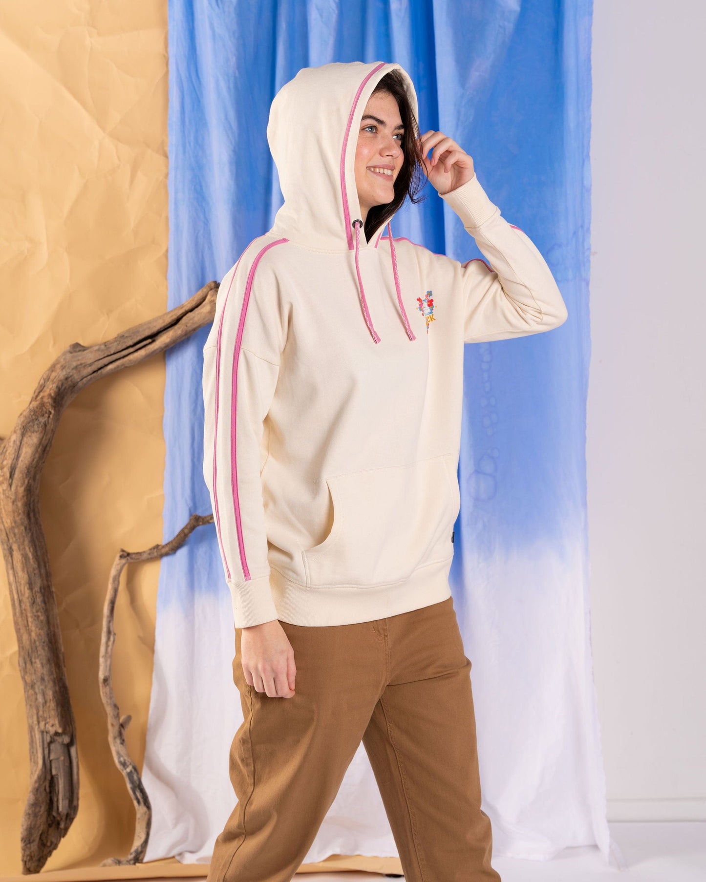 A person in a cozy Saltrock Highway Women's Pop Hoodie in cream and brown pants poses gracefully against a blue and beige backdrop with a delicate branch.