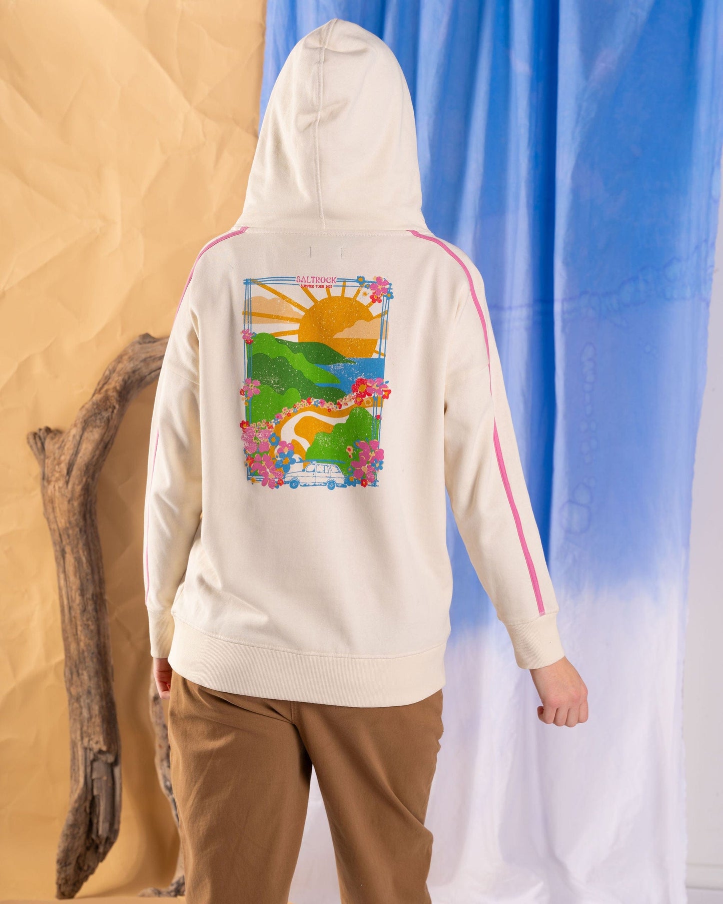 A woman wears the Saltrock Highway Women's Pop Hoodie in cream, featuring a vibrant landscape print with a sun, hills, and flowers on her back. She poses before a draped fabric backdrop with a wooden branch beside her.