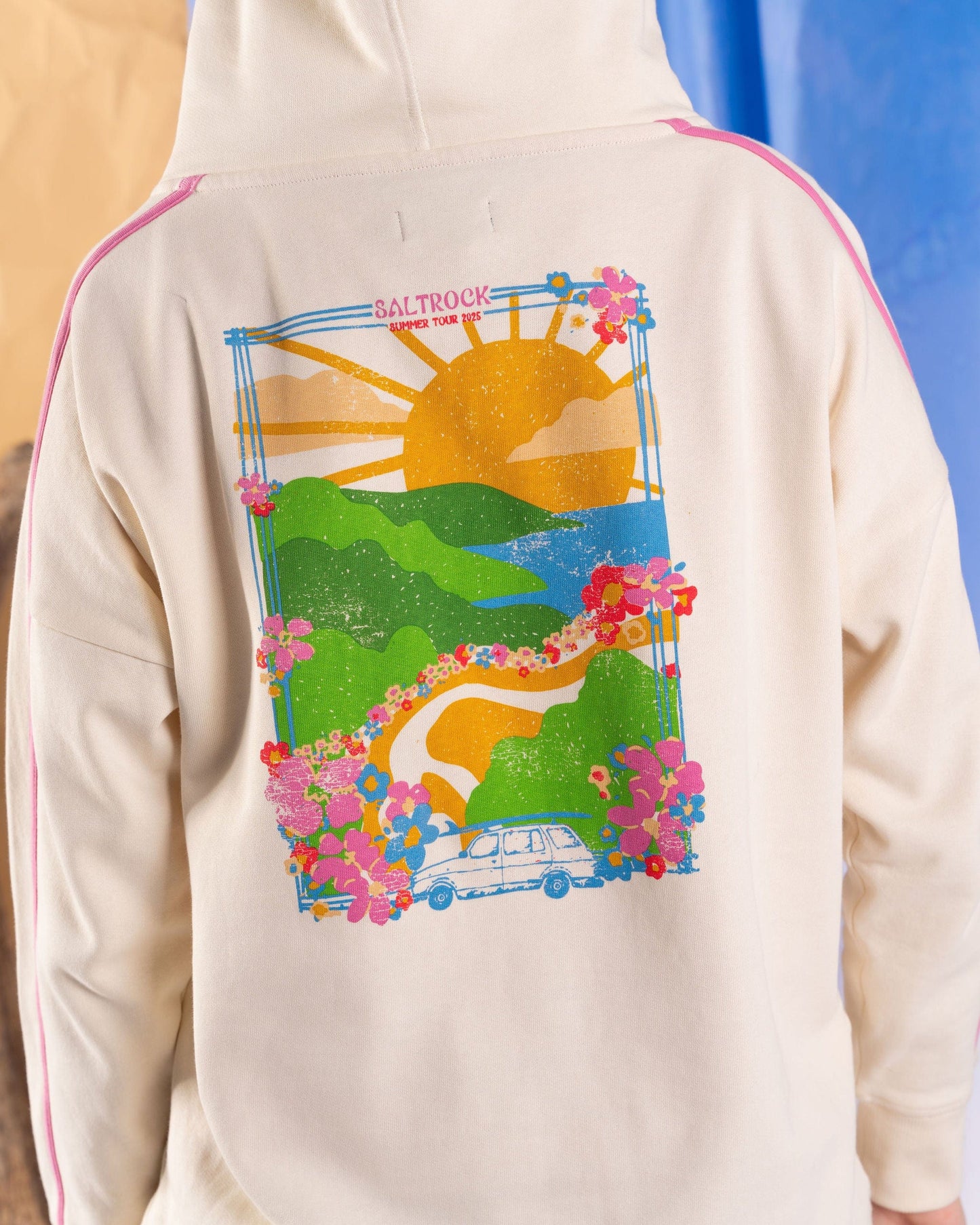 A person is wearing the Saltrock Highway Women's Pop Hoodie in cream, crafted from soft cotton with a vibrant back graphic depicting a scenic landscape including a sun, hills, flowers, and a car.