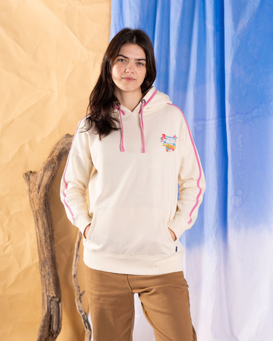 A person in a Saltrock Highway Women's Pop Hoodie - Cream and tan pants stands against a beige and blue backdrop, beside driftwood.